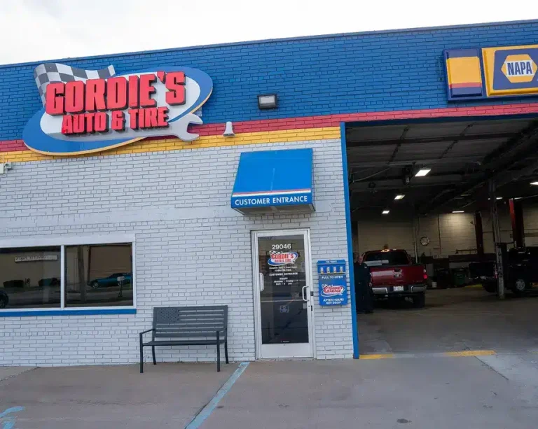 Exterior of Gordies Auto & Tire shop with a blue and white brick facade, a bench by the entrance, and a NAPA Auto Parts sign above the open garage bay.