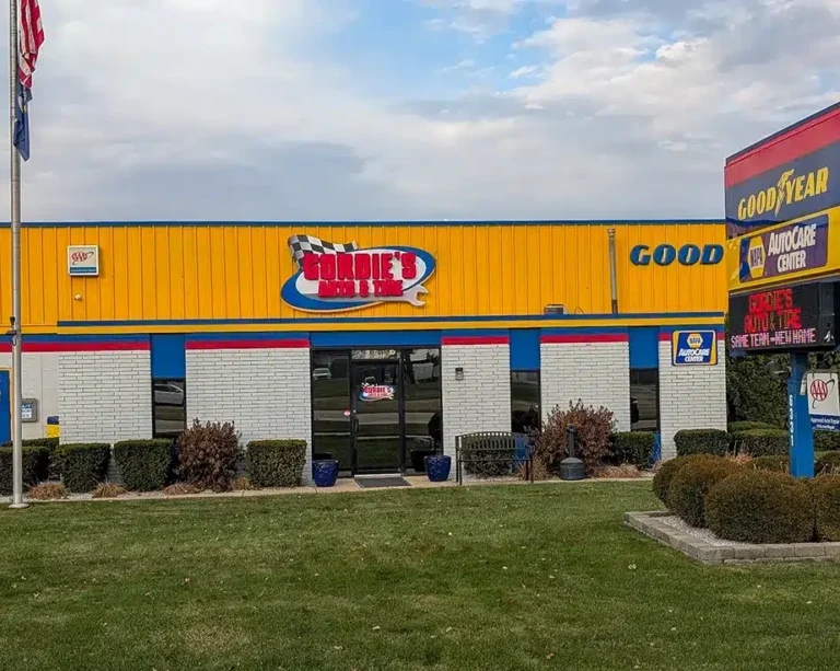 A yellow and white auto repair shop labeled Gordies Auto Tire with a Goodyear sign, glass doors, trimmed bushes, and a grassy lawn in front under a partly cloudy sky.
