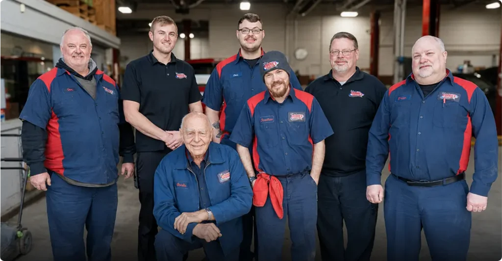 Seven men, most wearing matching blue and red work uniforms, pose together and smile inside a brightly lit auto repair shop. Tools and equipment are visible in the background.