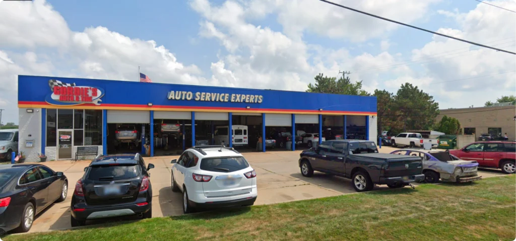 A blue auto repair shop with six open garage bays, cars parked in front, and a sign reading Auto Service Experts on the building. Trees, grass, and a partly cloudy sky are in the background.