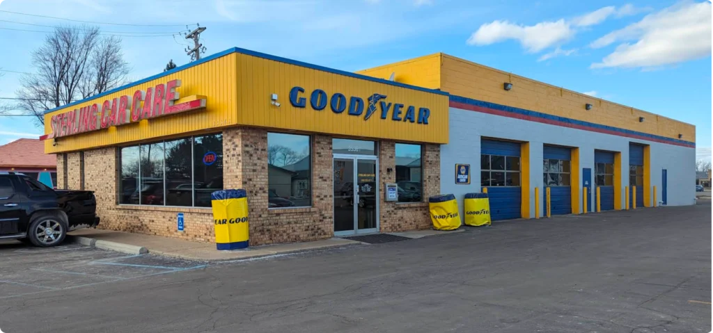 A Goodyear tire and auto care shop with large windows, a yellow sign, and multiple garage bays. Yellow tire covers and a blue sky are visible outside the building.