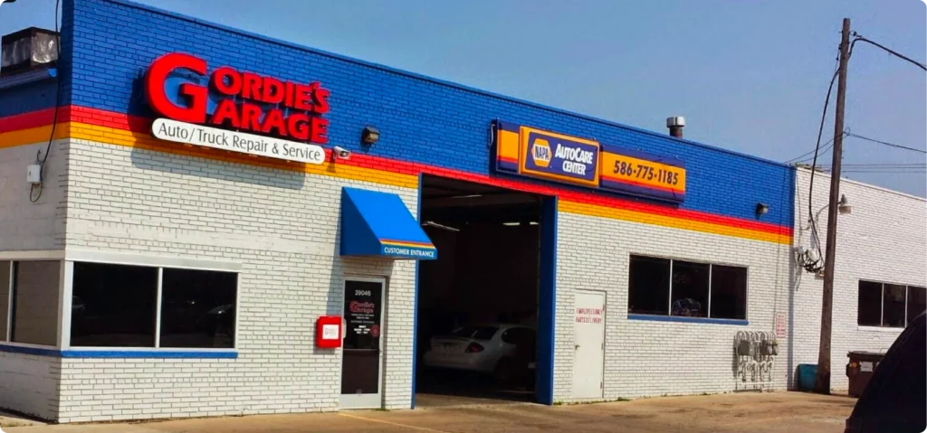Front view of Gordies Garage, an auto and truck repair shop with a blue, red, and yellow facade. A white car is partially visible inside the open service bay.