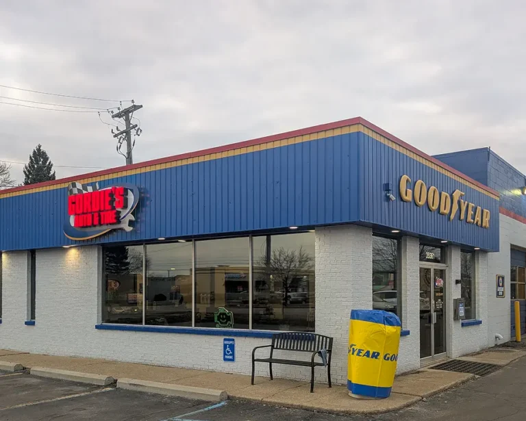 A Goodyear auto shop with Gordies Auto & Tire signage, large front windows, a bench, a blue-and-yellow trash can, and a handicap parking sign out front under a cloudy sky.