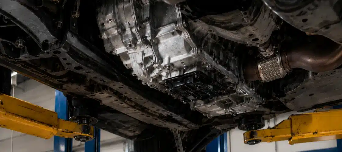 Close-up view of a cars undercarriage on a lift, showing the transmission, exhaust pipe, suspension parts, and metal framework in an automotive workshop.