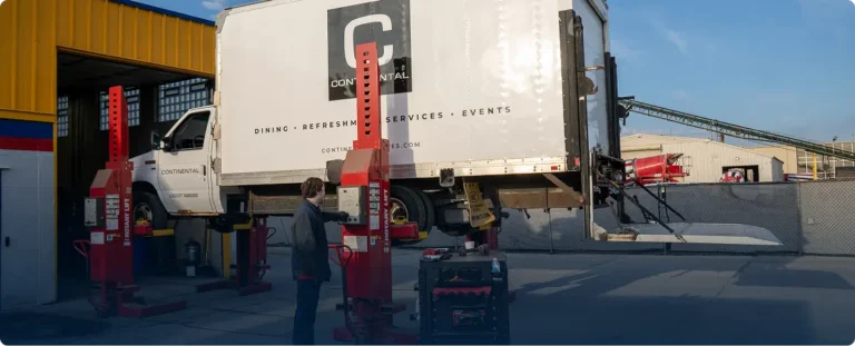 A worker operates a vehicle lift raising a large white Continental service truck outside Gordie’s Auto & Tire, with equipment and machinery visible in the background.